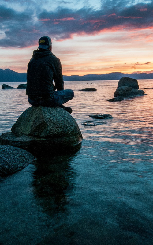 A man sits on a rock amidst a vast expanse of water beneath a sky in breathtaking colors from orange to pink, blue, and violet, showcasing what a balanced and fulfilling life may look like.